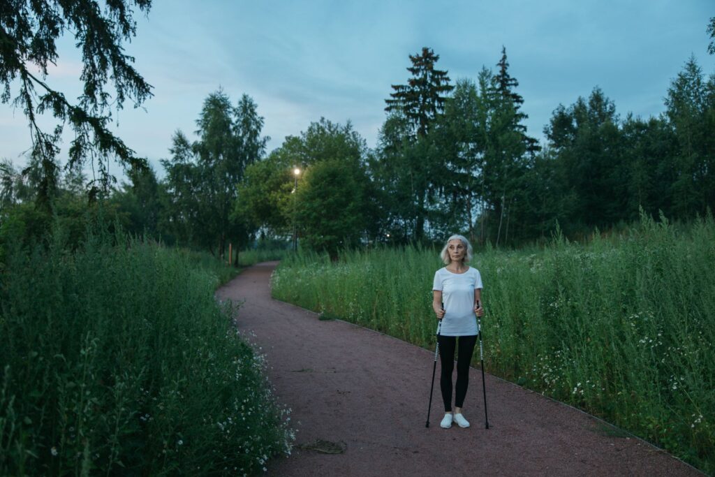 Kvinde med kort, lyst hår og hvid T-shirt står med stavgangsstave på en sti mellem højt, grønt græs og træer
