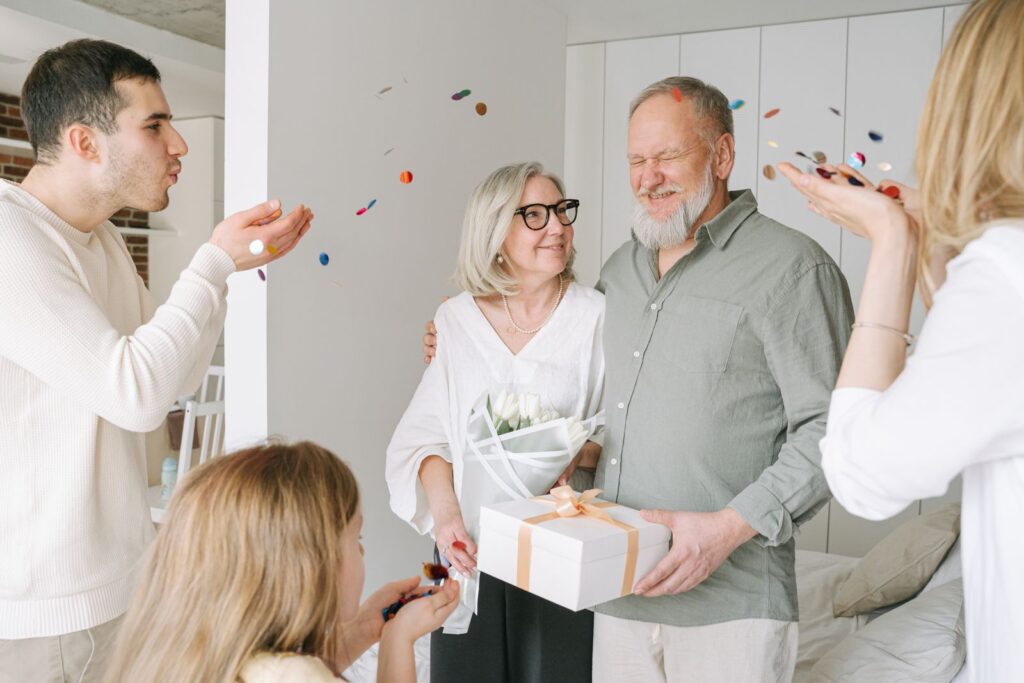 Glad ældre par med gaveæske og tulipaner, mens familien blæser konfetti om dem i et lyst hjem