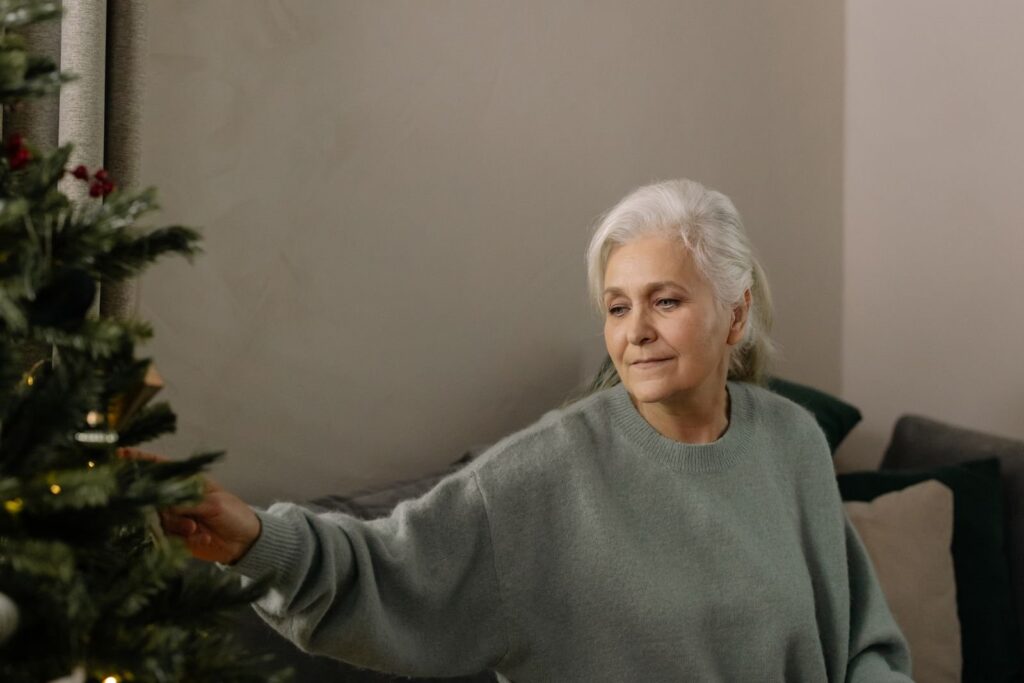 Kvinde med hvidt hår og grå sweater pynter et juletræ i en stue med en krans på væggen