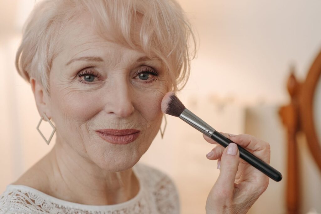 Kvinde med kort lyst hår og øreringe smiler og fører en makeuppensel op mod kindbenet tæt ved øjet