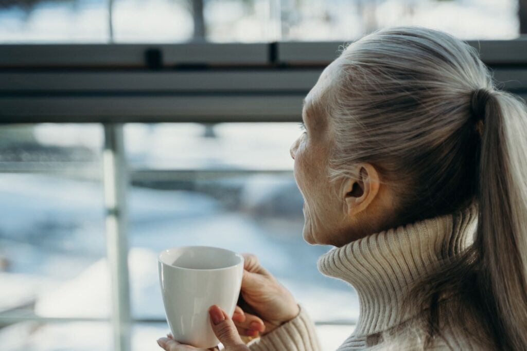Kvinde med langt gråt hår i hestehale og cremefarvet rullekravesweater holder en kop og kigger ud ad et vindue på en vinterdag